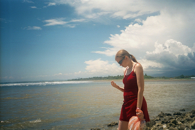 Adrienne standing on a beach looking down at her hand and looking mildly angry. 